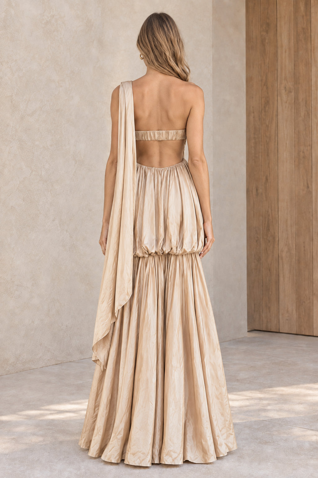 Back view of woman wearing champagne maxi dress highlighting cut-out back and draped shoulder detail